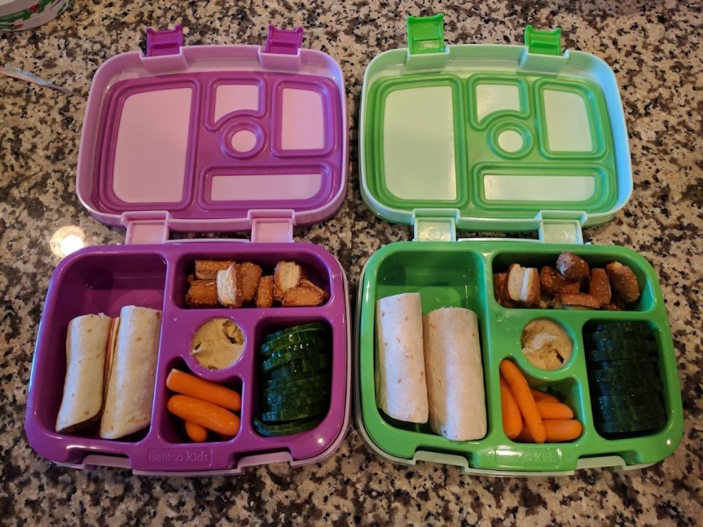 two open bento lunch boxes filled with carrots, cumbers hummus, pretzels and sandwich wraps